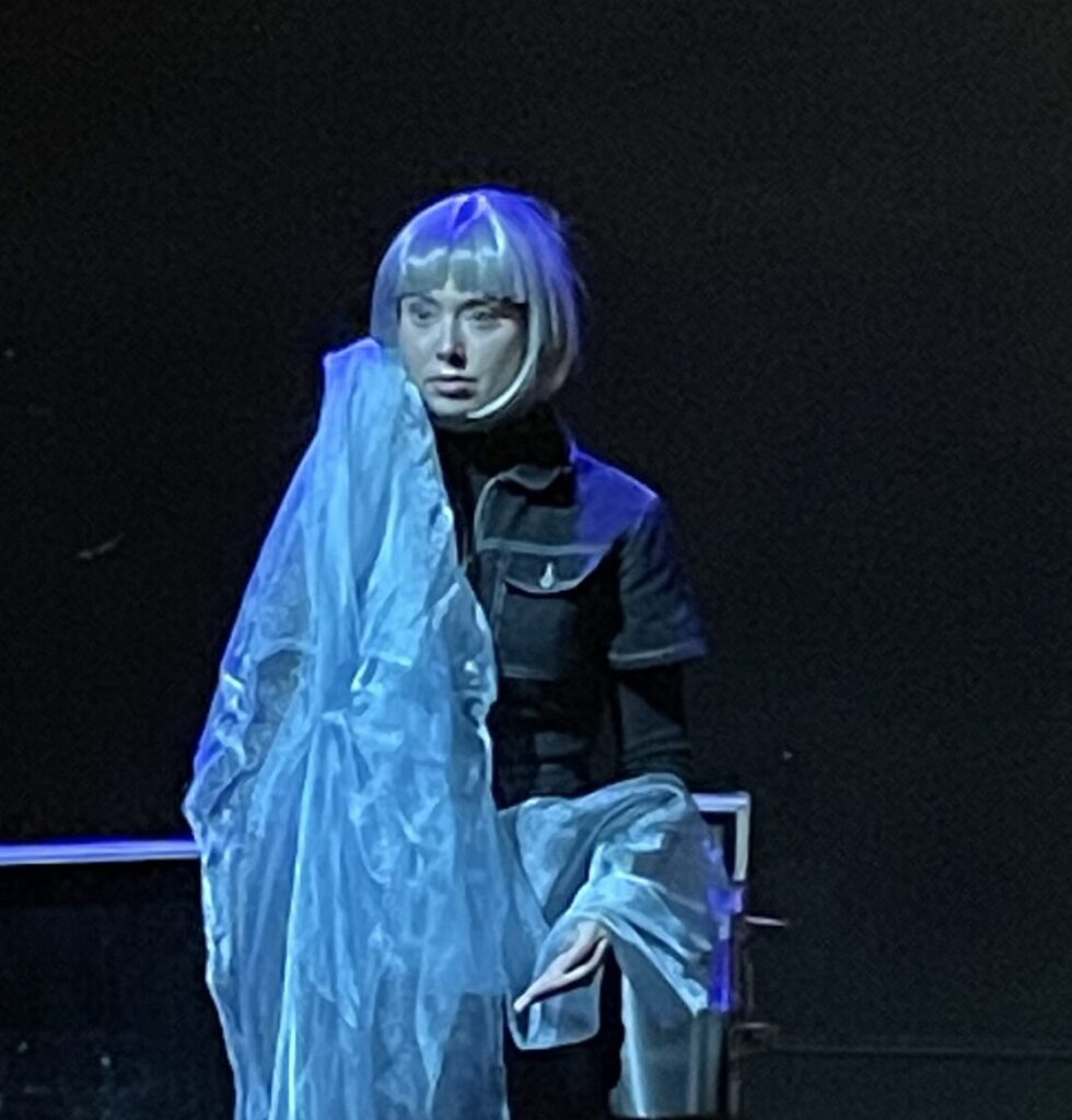 A female actor wearing all black clothing and wearing a silver wig stands on a black stage with a black backdrop. She clutches a long piece of light blue cloth to her body.