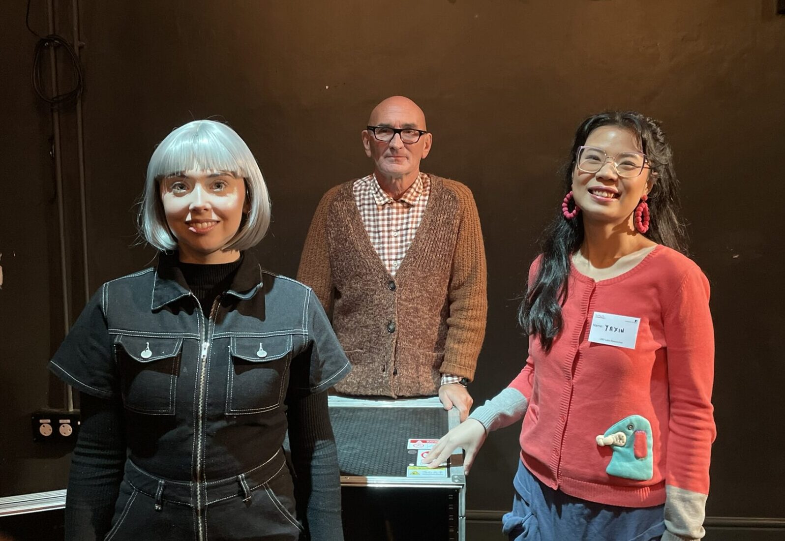 Three people appear in the photograph standing in a theatre with black back drop. A female actor stands on the left dressed in black and wearing a silver wig. A male wearing glasses, a checked shirt and brown cardigan stands in the centre. A female actor wearing glasses, a bright pink cardigan and pink hoop earrings stands at the right. She wears a name tag labelled Yaxin.