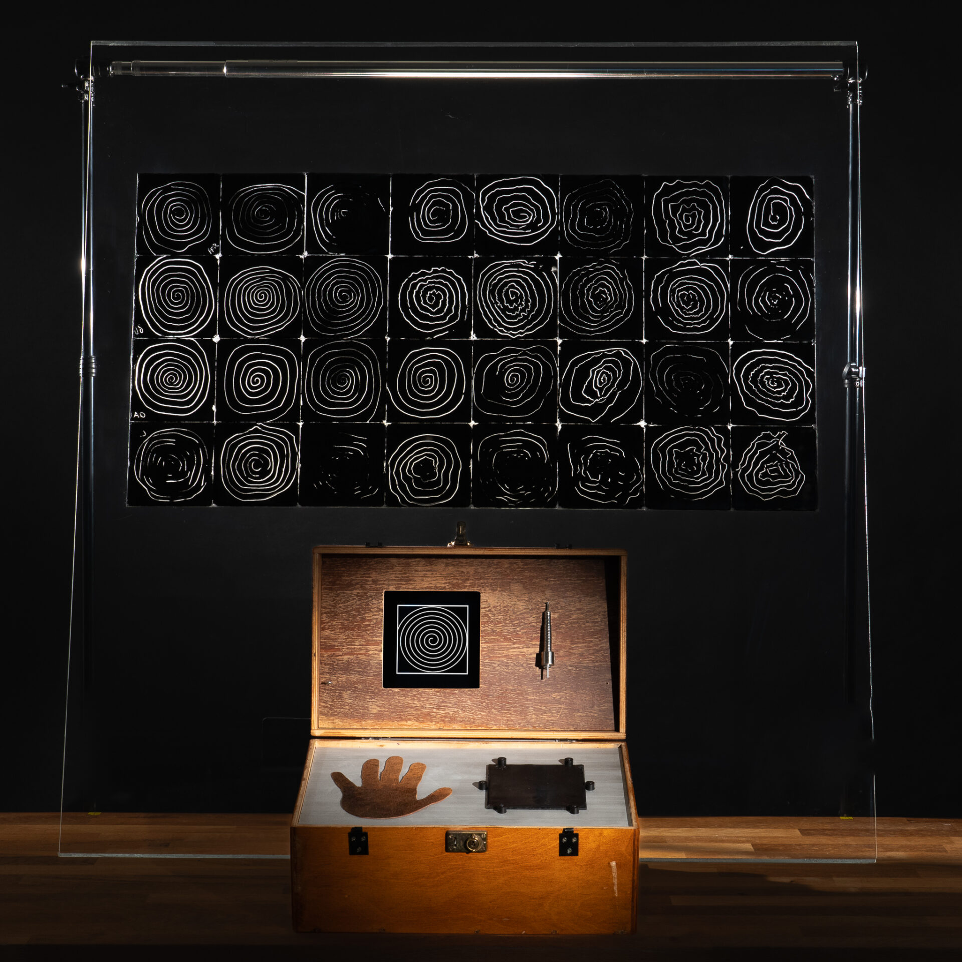 On a black wall is a 9 x 4 grid of black tiles with white spiral engravings on them. In front of this is an open wooden box, housing a small stylus, hand print, and space to fix sample tile for engraving.