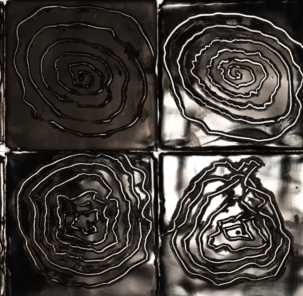 Close up of four black tiles positioned next to one another to form a square. Each has an engraved white spiral on it, drawn unevenly.