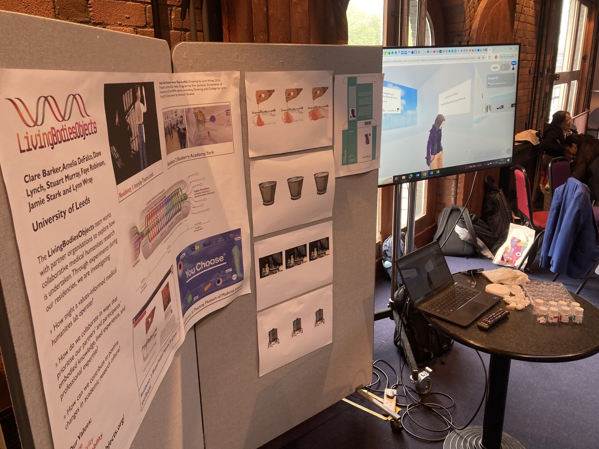 Two grey poster boards in a meeting room. Attached to them are a poster about the LBO project and a number of drawings. Next to the poster boards is a round table with a laptop upon it, linked to a display screen showing a VR space.